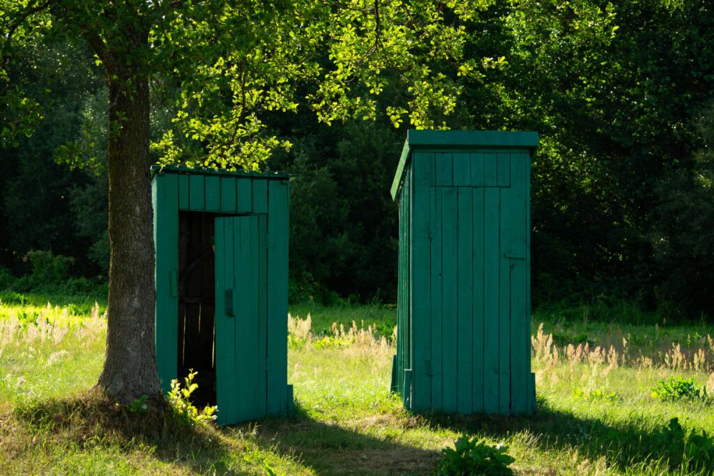Two green wooden huts in a sunny rural setting surrounded by lush greenery.