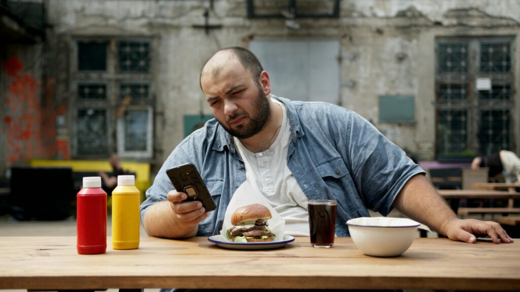 Bald bearded man sitting outdoors holding smartphone with burger and condiments.