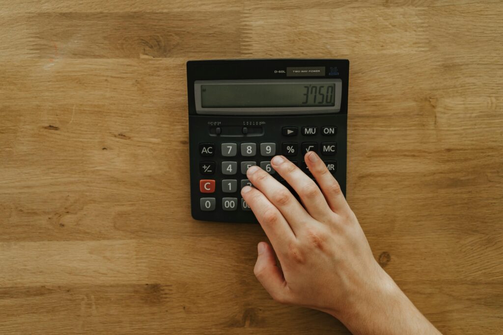 A person's hand pressing keys on a calculator displaying 3750 on a wooden table, top view.